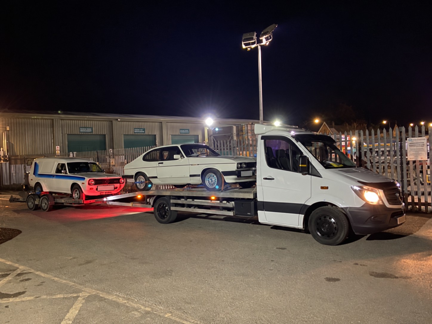 Two classic cars carried on a recovery truck and trailer at night