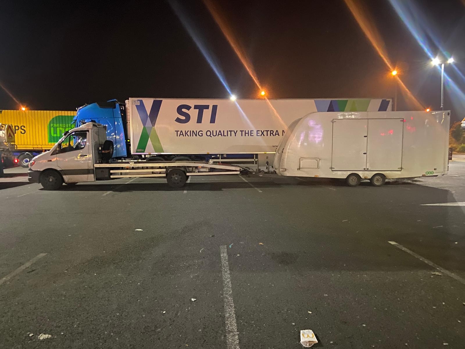 Flatbed recovery truck towing an enclosed white trailer at night